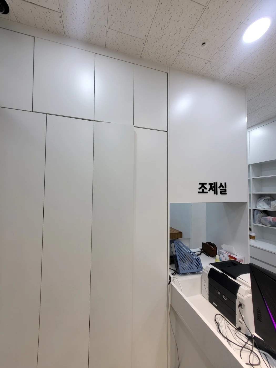 카운터 옆의 문이 달린 흰색 수납장과 조제실 표지가 있는 벽